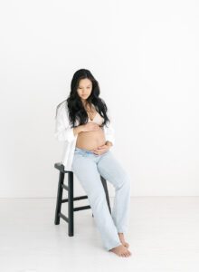 pregnancy photo of mom in jeans and white button down on a black stool