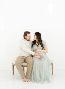 maternity photo of women in green ASOS dress sitting on bench with husband in natural light photography studio