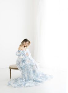 mom sitting on a bench in natural light studio for maternity photos in blue and white floral puffy dress