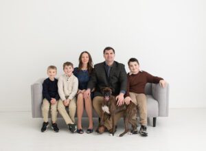 Family photos with 3 boys and a boxer dog in studio on a couch against white wall.