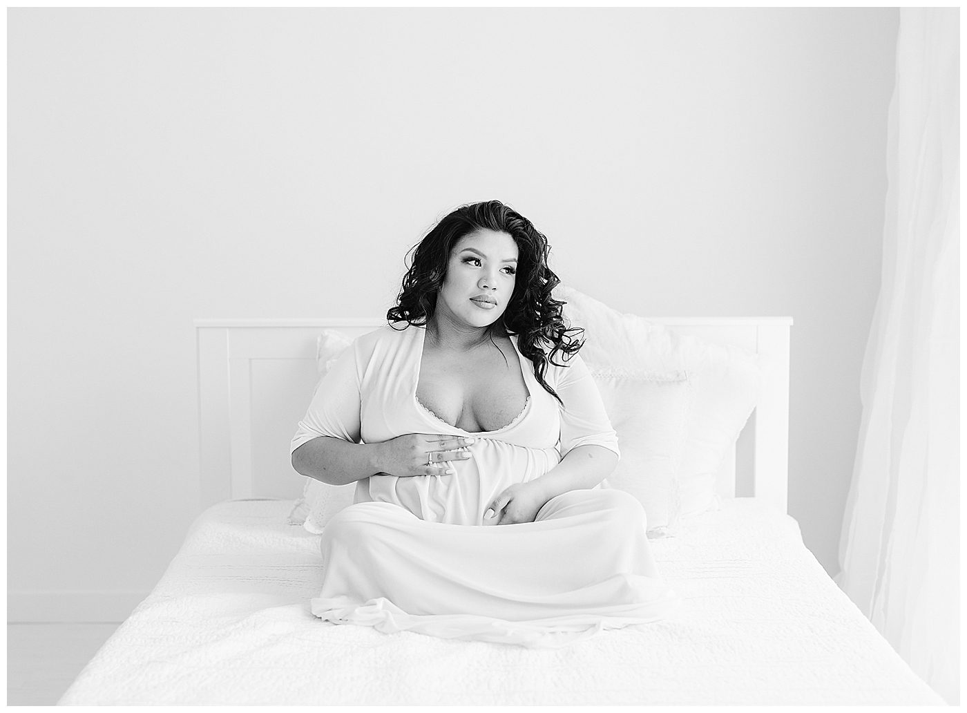 black and white maternity photo of mom in long white dress sitting on bed