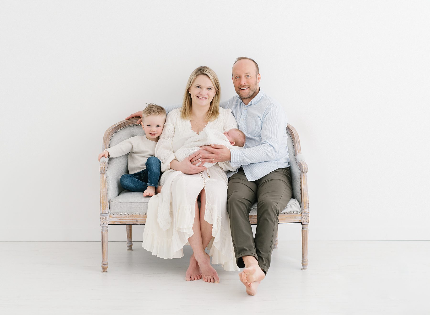 family with toddler and newborn sitting on grey loveseat along a white wall