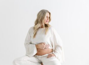 pregnant mom with linen top and pants sitting on stool with hand on belly, looking off to the side against a white wall in a Minneapolis natural light photography studio