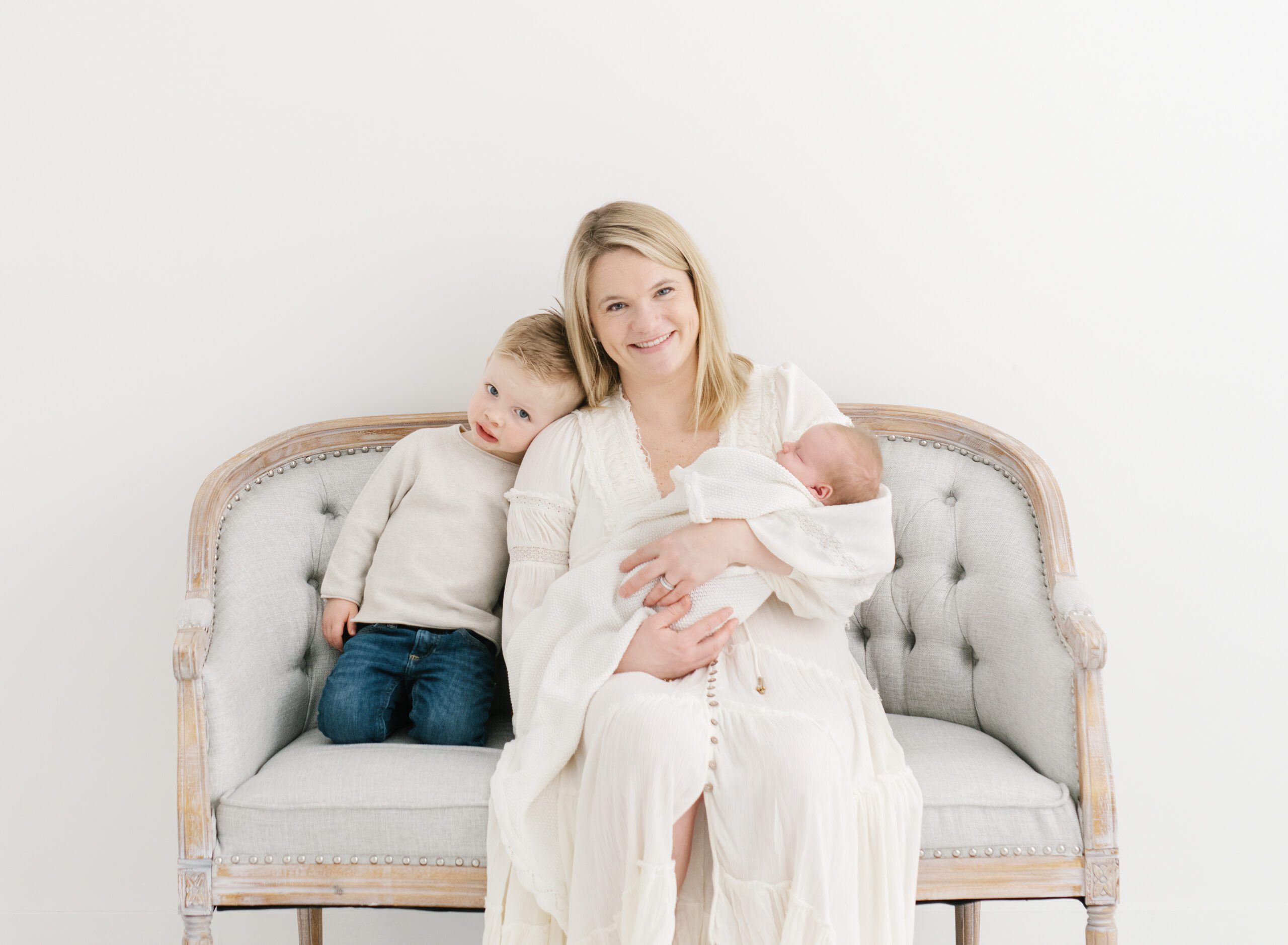 mom in long white Free People dress sitting on grey couch with her a newborn and a toddler son