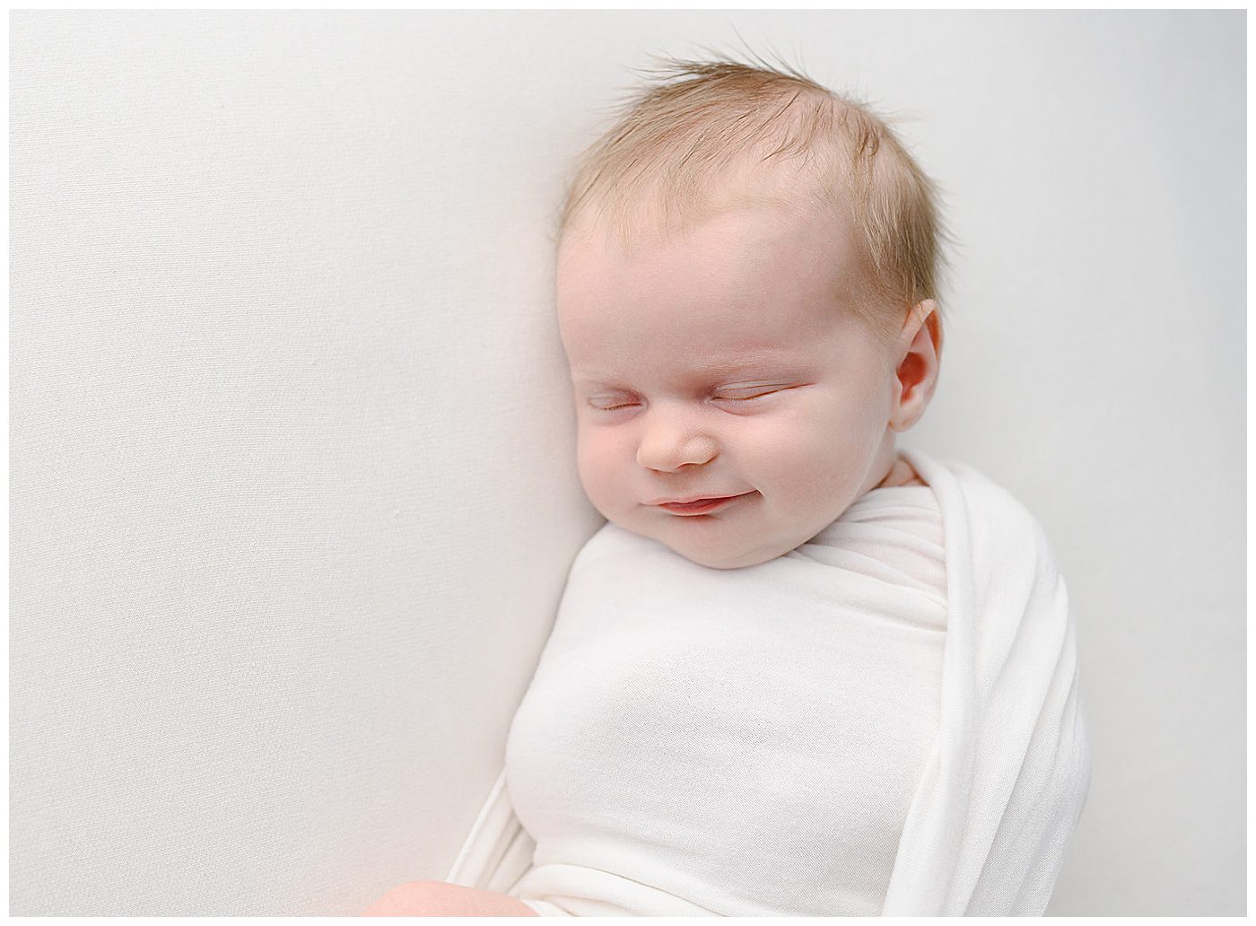 Edina newborn photographer photographing newborn baby swaddled in white on white backdrop, with baby smiling