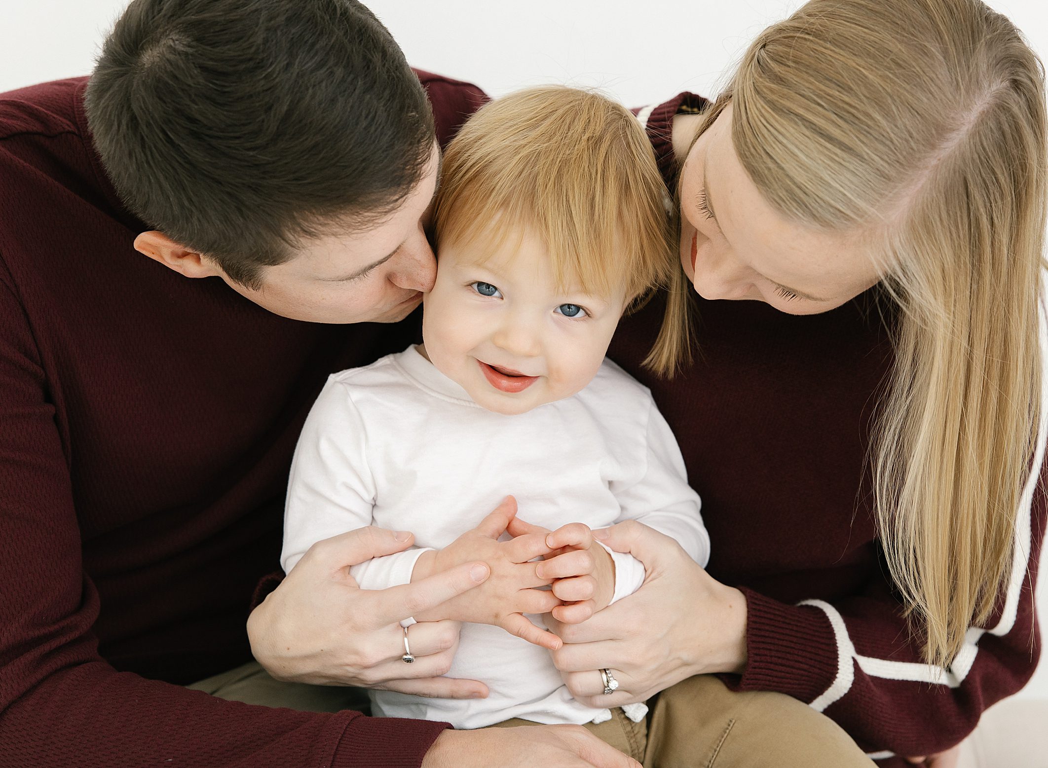 Mom and dad in maroon shirts holding one year old baby boy in white shirt between them
