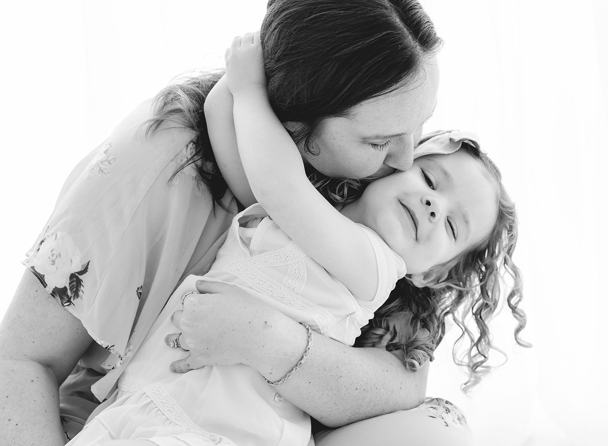 black and white backlit photo of mom kissing toddler daughter, who is hanging on to her neck while sitting in her lap